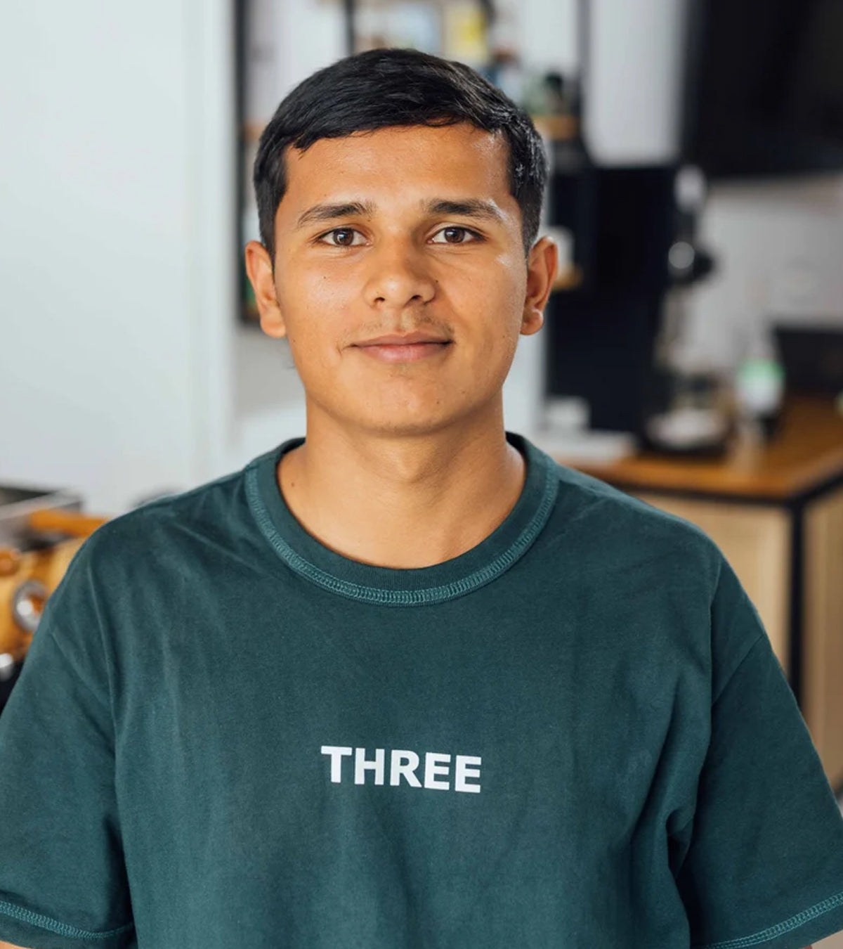 Marlon Bolaños wearing a dark green crewneck t-shirt with the word 'THREE' printed in white on the chest, photographed in a softly blurred indoor setting.