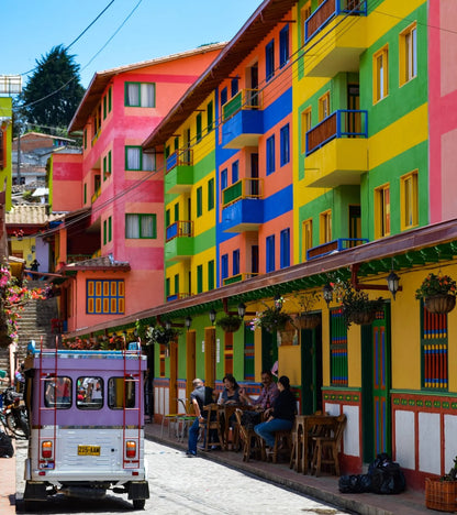 Colorful colonial-style buildings painted in bright stripes of pink, orange, yellow, green, blue, and white along a cobblestone street, with outdoor dining seating and a small white vehicle in the foreground.
